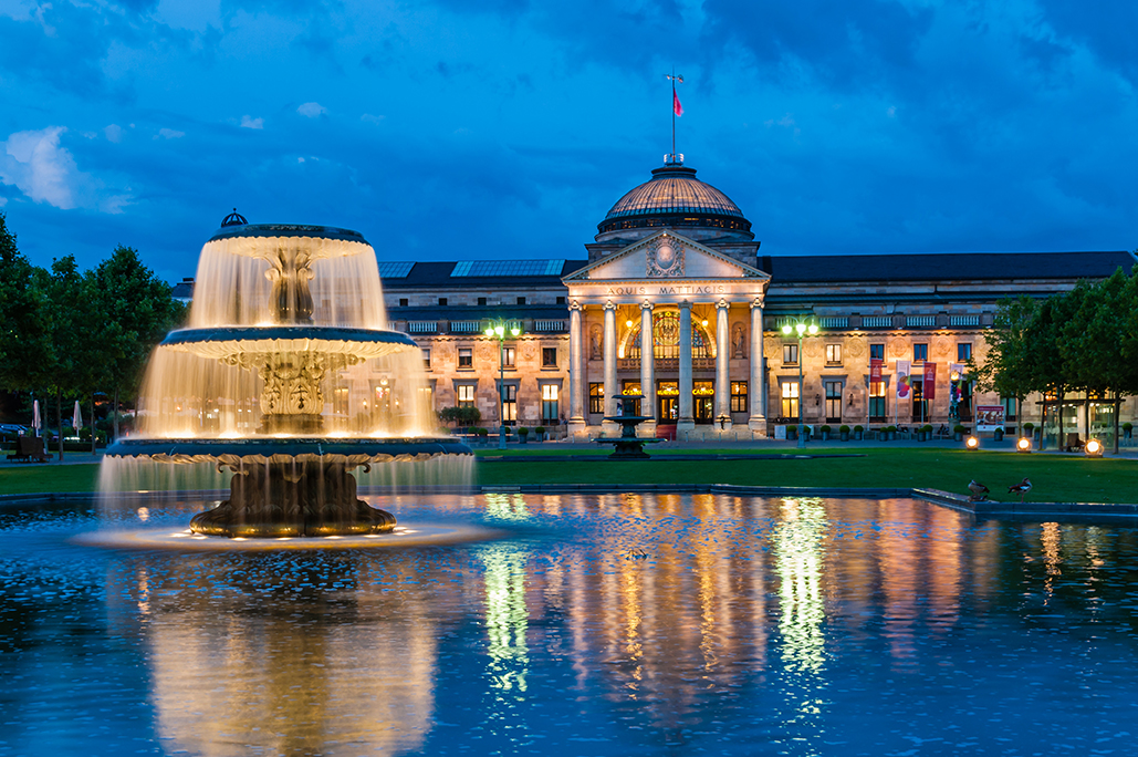 Bild des Wiesbadener Kurhaus bei Nacht. Das Gebäude ist beleuchtet, im Vordergrund befindet sich ein ebenfalls beleuchteter Brunnen, in dessen Wasser sich das Kurhaus widerspiegelt.